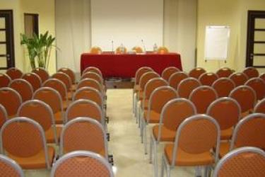 Sala konferencyjna z rzędami pustych krzeseł i stołem z mikrofonami.