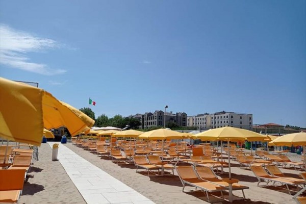 Plaża z rzędami pomarańczowych leżaków i żółtymi parasolami, w tle budynki i włoska flaga.