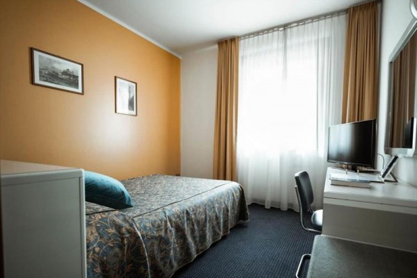 Prosty pokój hotelowy z łóżkiem, telewizorem i oknem z brązowymi zasłonami.