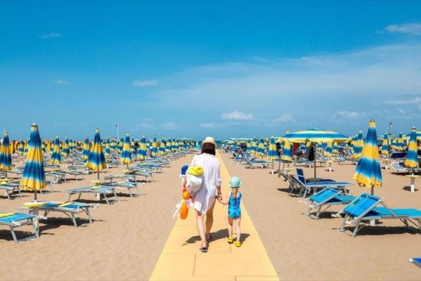 Kobieta z dzieckiem idzie po piaszczystej plaży między leżakami i parasolami.