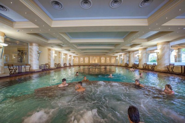 Ludzie relaksujący się w krytym basenie z jacuzzi i kolumnami po bokach.