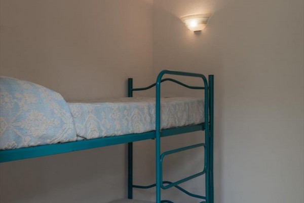 Metalowe łóżko piętrowe z niebieskimi materacami obok białej ściany z lampą.