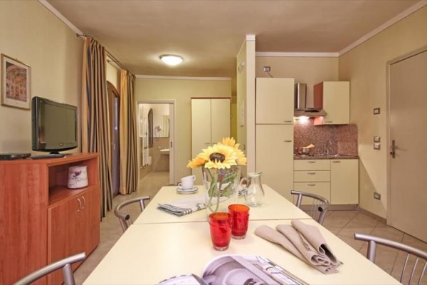 Kuchnia z jadalnym stołem, kwiatami i jasnymi meblami w apartamencie z otwartymi drzwiami do łazienki.
