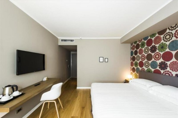 Nowoczesny pokój hotelowy z podwójnym łóżkiem, telewizorem, biurkiem i kolorową tapetą na jednej ścianie.