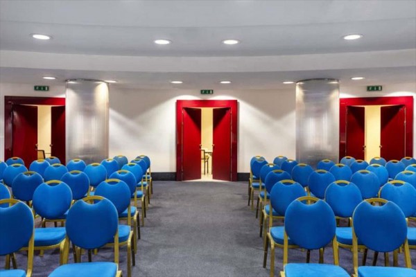 Sala konferencyjna z niebieskimi krzesłami i czerwonymi drzwiami z tyłu.