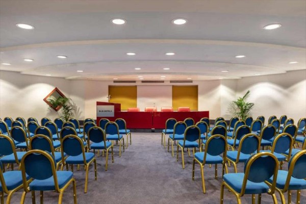 Sala konferencyjna z rzędami niebieskich krzeseł i czerwonym stołem z przodu.