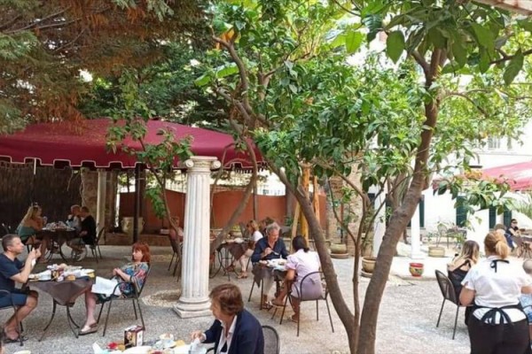 Ludzie siedzą i jedzą w ogrodowej restauracji pod drzewami i czerwonymi parasolami.
