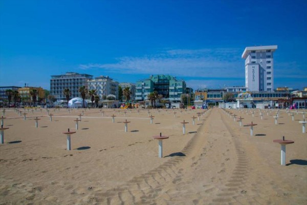 Piaszczysta plaża z pustymi stojakami na parasole i w tle miejskie budynki.