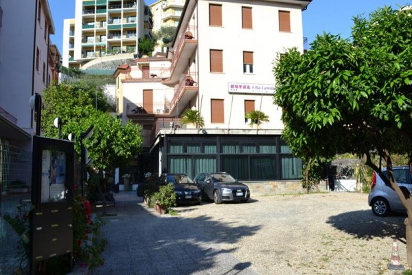 Budynek hotelu otoczony drzewami i zaparkowanymi samochodami.