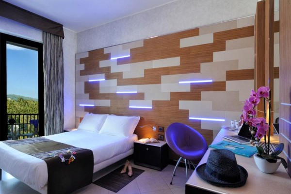 Nowoczesny pokój hotelowy z podwójnym łóżkiem, fioletowym fotelem i dekoracyjnymi światłami na ścianie.