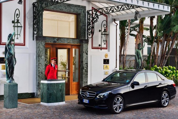Portier w czerwonym mundurze stoi przy wejściu do budynku obok czarnego samochodu Mercedes-Benz.