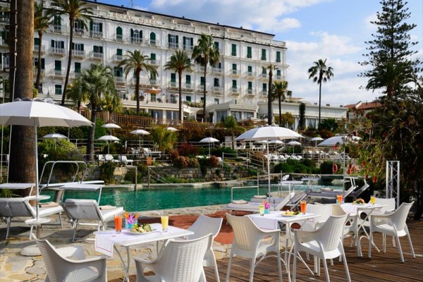 Hotel z basenem, otoczony palmami i parasolami, z zewnętrznymi stolikami i krzesłami.