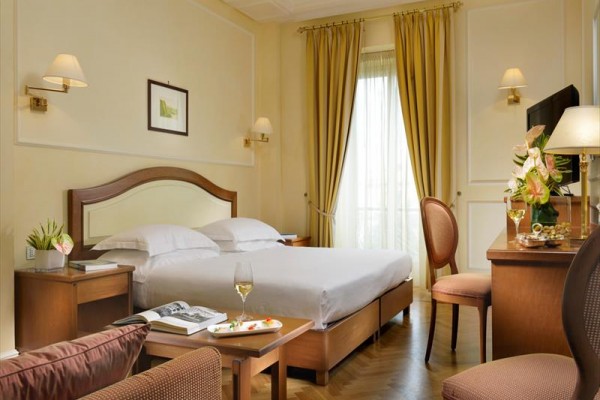 Elegancko urządzony pokój hotelowy z podwójnym łóżkiem, fotelami, biurkiem i złotymi zasłonami.