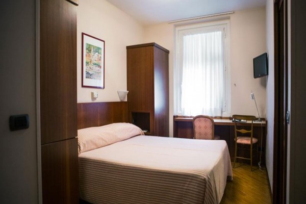 Prosto urządzony pokój hotelowy z łóżkiem, stolikiem, krzesłem i telewizorem na ścianie.