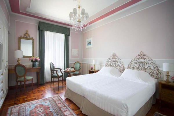 Elegancko urządzony pokój hotelowy z podwójnym łóżkiem, fotelami i stołem, w pastelowych kolorach.