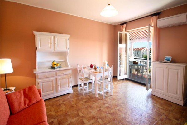 Jasny salon z jadalnianym stołem, białymi meblami i otwartymi drzwiami na balkon.