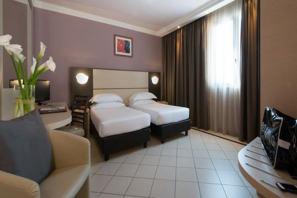 Pokój hotelowy z dwoma oddzielnymi łóżkami, fotelem i bagażem obok stolika.
