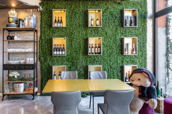 Restauracja z sztuczną zieloną ścianą ozdobioną butelkami wina i stołem z pluszowym misiem.