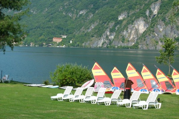 Białe plastikowe leżaki na trawie na pierwszym planie, za nimi jezioro z kolorowymi żaglami windsurfingów i zalesione góry.