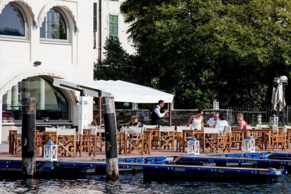 Taras restauracji z drewnianymi stołami i krzesłami nad wodą, gdzie kelner obsługuje gości.
