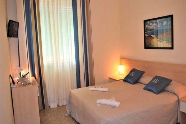 Mały pokój hotelowy z łóżkiem małżeńskim, zasłonami i obrazem na jasnoniebieskiej ścianie.  