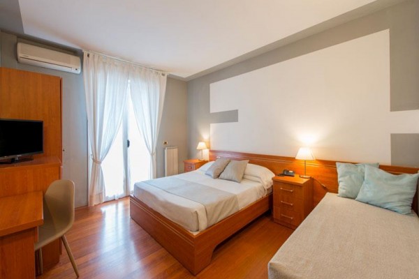 Pokój hotelowy z łóżkiem małżeńskim, pojedynczym łóżkiem, biurkiem, telewizorem i lampkami na stolikach nocnych.
