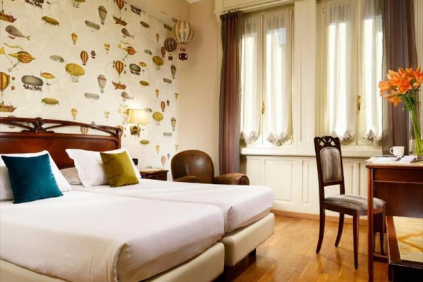 Pokój hotelowy z dwoma pojedynczymi łóżkami, tapetą z balonami na gorące powietrze i oknami z długimi zasłonami.