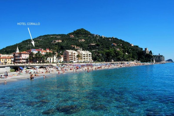 Plaża z ludźmi i budynkami na wybrzeżu pod zalesionym wzgórzem.