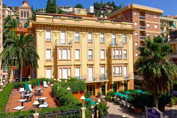 Żółty hotel z tarasem i palmami na tle miasta.