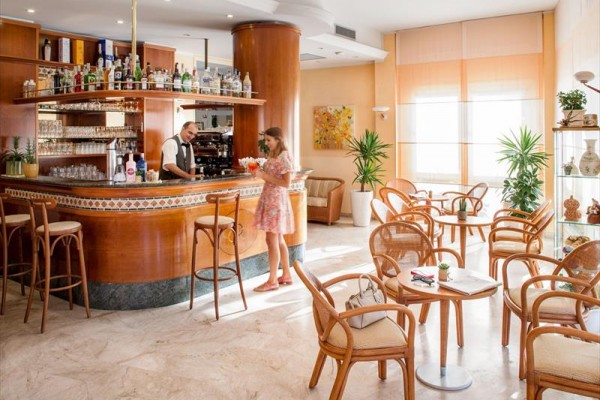 Kobieta zamawia drinka przy barze w hotelowym lobby z rattanowymi krzesłami i roślinami.
