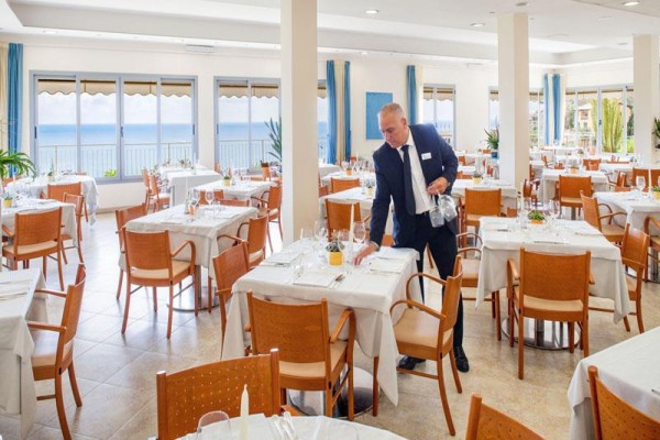 Kelner w garniturze poprawia stół w jasnej restauracji z widokiem na morze.
