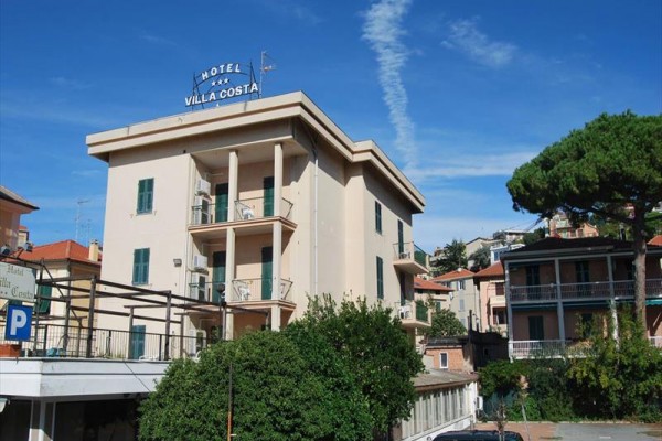 Hotel Villa Costa to czteropiętrowy budynek otoczony zielenią i innymi budynkami.