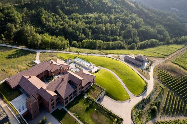 Widok na nowoczesny budynek otoczony zielonymi łąkami i winnicami w pagórkowatym, zalesionym krajobrazie.