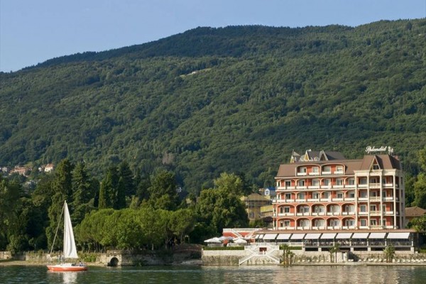 Budynek hotelu nad jeziorem z żaglówką na wodzie i zalesionym wzgórzem w tle.