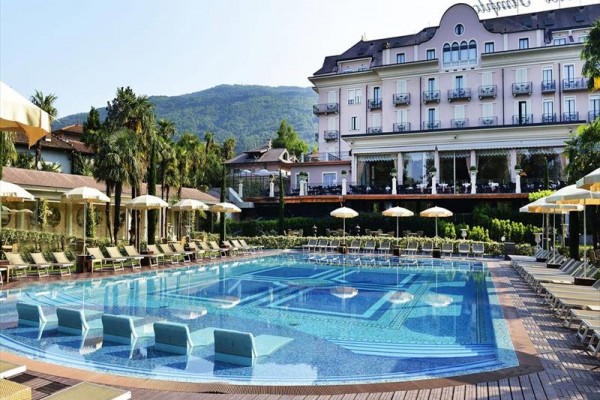 Duży hotel z basenem otoczonym leżakami i parasolami, w tle zielone wzgórza.