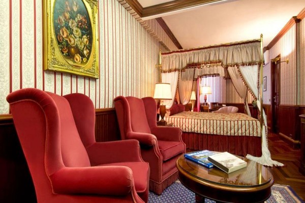 Luksusowy pokój hotelowy z łóżkiem z baldachimem, dwoma czerwonymi fotelami i obrazem kwiatów na ścianie.