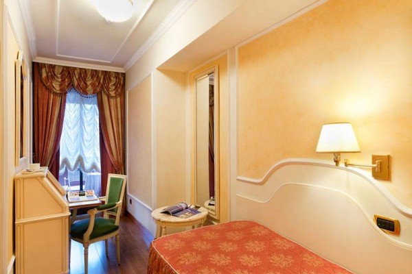 Wąski pokój hotelowy z pojedynczym łóżkiem, biurkiem i lampą na ścianie.