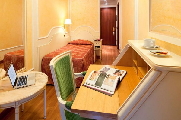 Zadany pokój hotelowy z pojedynczym łóżkiem, stołem z laptopem i otwartą książką.