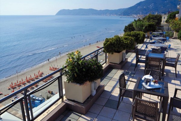 Taras z widokiem na morze, z zewnętrznymi miejscami do siedzenia i kwiatami, obok plaży z leżakami.