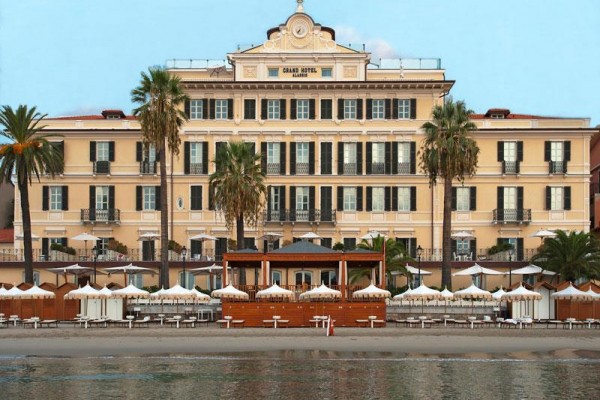 Duży hotel przy plaży z palmami i tarasem z białymi parasolami.