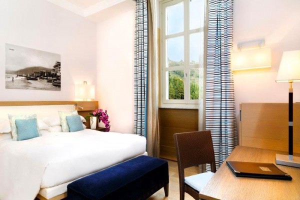 Nowoczesny pokój hotelowy z dużym łóżkiem, biurkiem i oknem z widokiem na zieleń.
