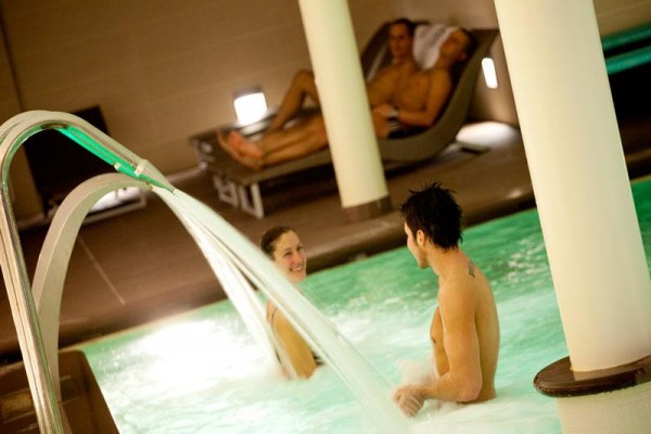 Dwie osoby śmieją się w jacuzzi, podczas gdy dwie inne relaksują się na leżakach w tle.