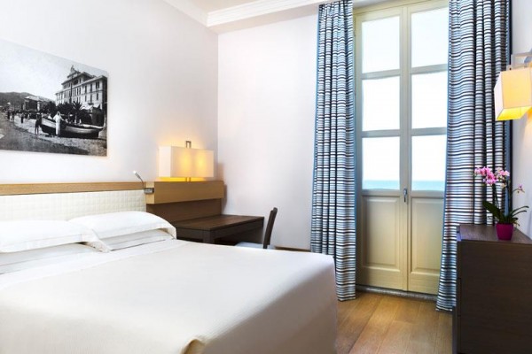 Pokój hotelowy z białym łóżkiem, biurkiem, zamkniętymi drzwiami na taras i zasłonami.