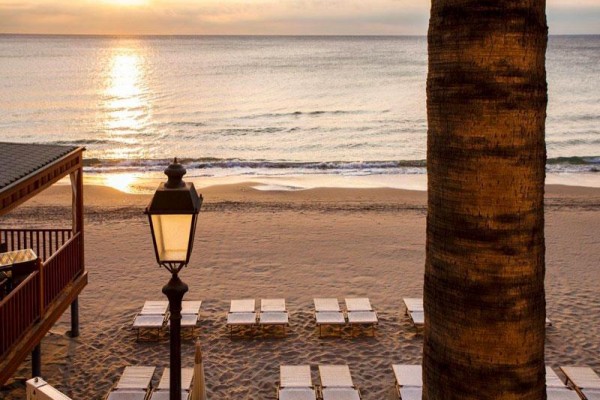 Piaszczysta plaża z leżakami o zachodzie słońca, na pierwszym planie palma i latarnia.