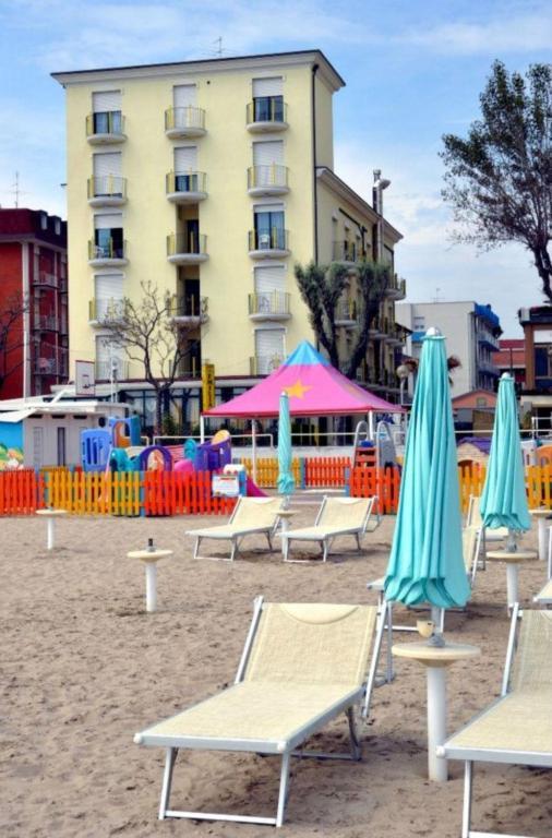 Leżaki i parasole na piaszczystej plaży przed kilkupiętrowym budynkiem i ogrodzonym placem zabaw.