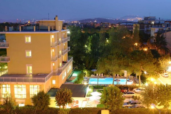 Wieczorny widok na oświetlony hotel z basenem otoczony drzewami i miejskim krajobrazem w tle.