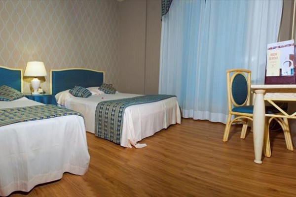 Dwa jednoosobowe łóżka z niebieskimi zagłówkami, stolik nocny z lampą, biurko i krzesło w hotelowym pokoju.