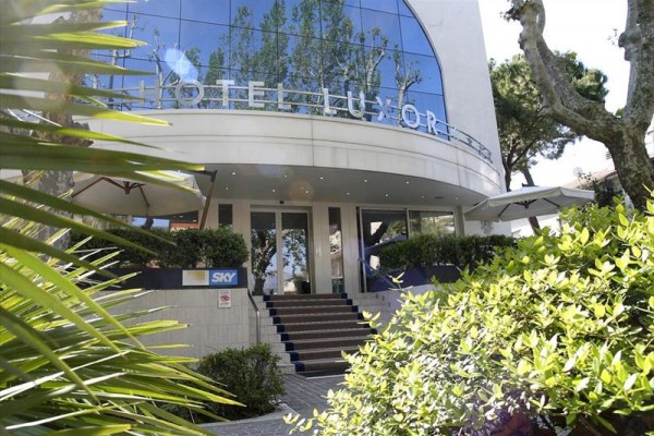 Wejście do hotelu Luxor z nowoczesną szklaną fasadą i schodami otoczonymi zielenią.