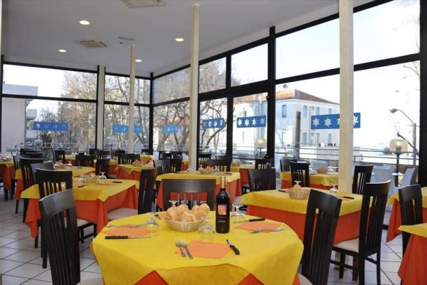 Restauracja z przeszklonymi ścianami, stoły z żółtymi obrusami i przygotowanymi sztućcami.