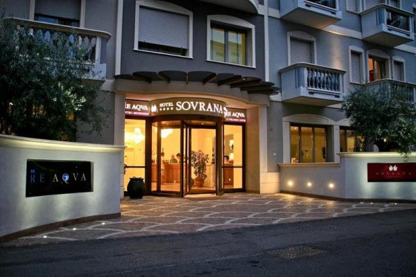 Główne wejście do hotelu Sovrana z oświetlonym logo i ozdobnymi roślinami.